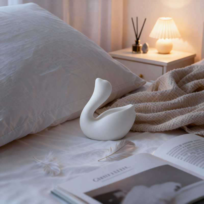 The elegant white swan massager resting on a bed, showcasing its discreet design in a private bedroom setting.